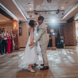 Pani-Migawka - Para młoda tańczy pierwszy taniec na parkiecie, otoczona gośćmi weselnymi. W tle ceglane kolumny i dekoracje sali balowej. Oświetlenie punktowe tworzy romantyczny nastrój.