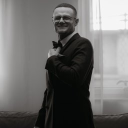 Pani-Migawka - Czarno-białe zdjęcie uśmiechniętego mężczyzny w garniturze i okularach, poprawiającego muchę. W tle okno i lampa, tworzące subtelne oświetlenie.