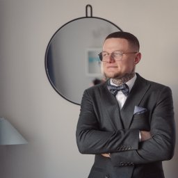 Pani-Migawka - Elegancki mężczyzna w garniturze i muszce, z tatuażami na szyi, stoi na tle lustra i lampy, spoglądając w bok. Ujęcie portretowe, stonowana kolorystyka.