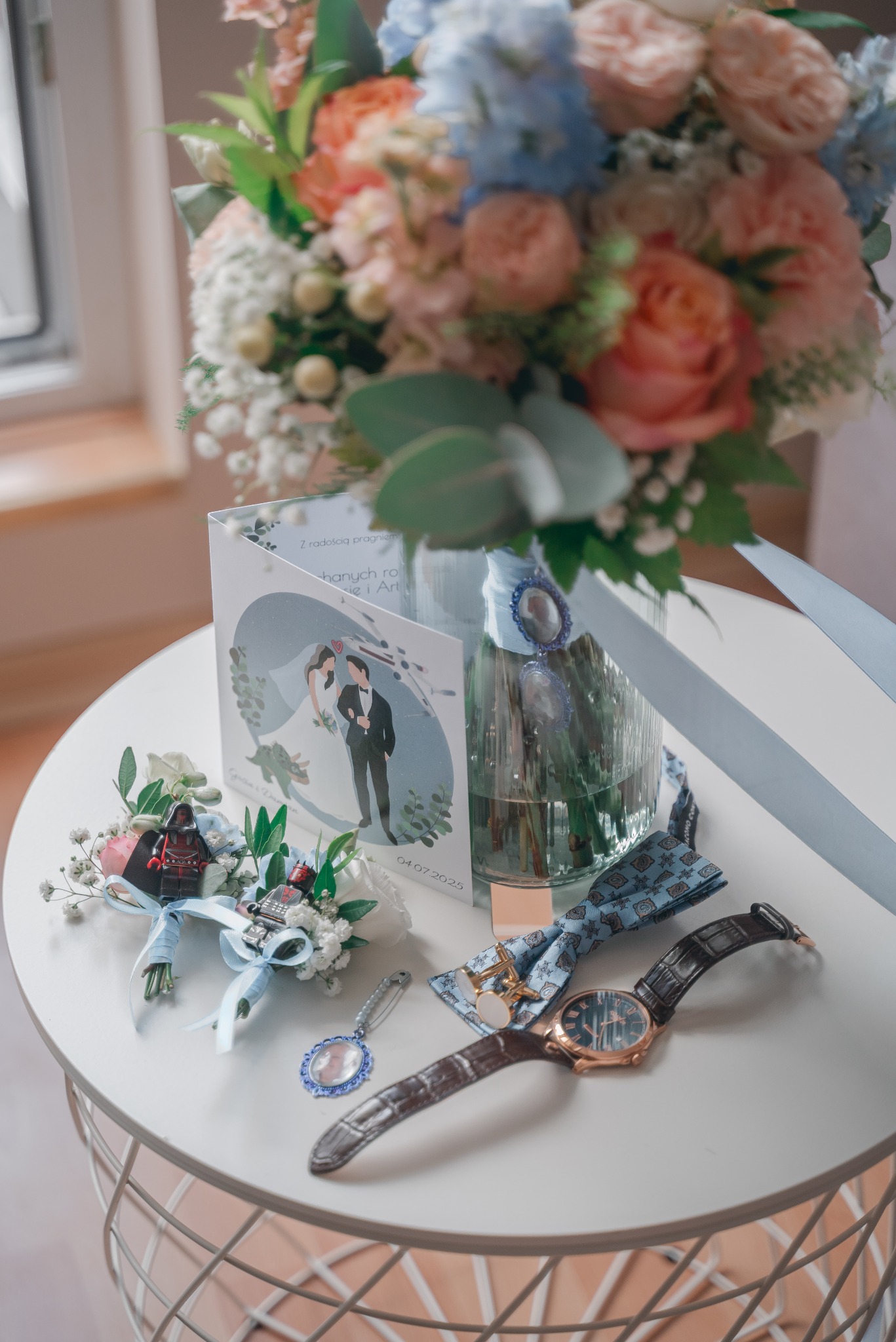 Bukiet ślubny w wazonie, zaproszenie z parą młodą, mucha, zegarek i butonierki z motywem Star Wars na białym stoliku. Detale ślubne w pastelowych kolorach.