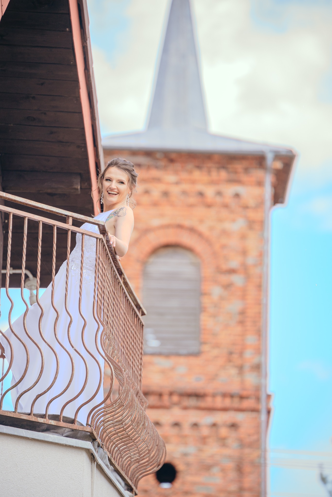 Uśmiechnięta panna młoda w białej sukni na balkonie z metalową balustradą, w tle ceglany kościół z wieżą. Jasne, naturalne światło.