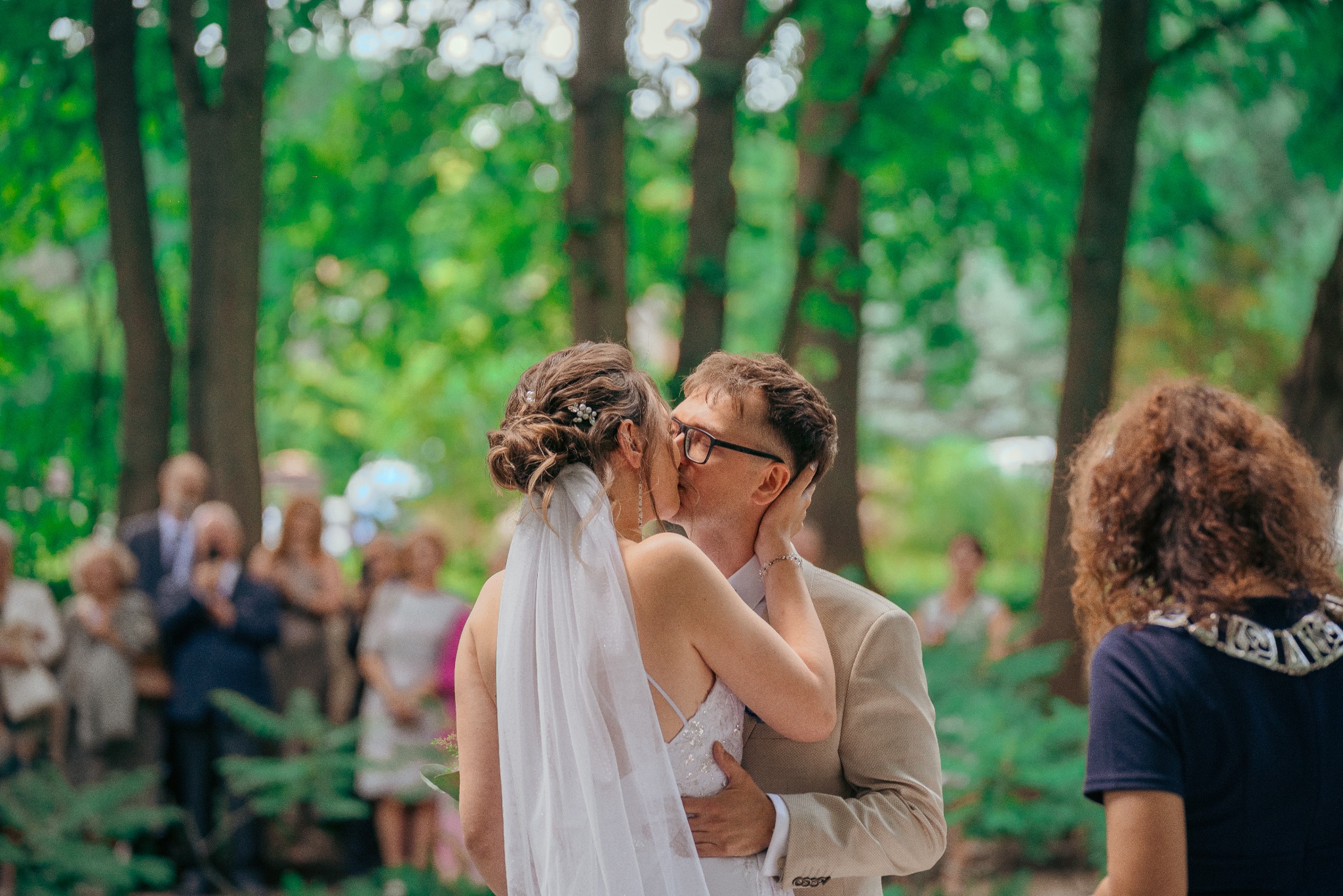 Pocałunek pary młodej w plenerze, panna młoda w sukni z odkrytymi plecami i welonem, pan młody w beżowym garniturze i okularach, goście w tle, naturalne światło.