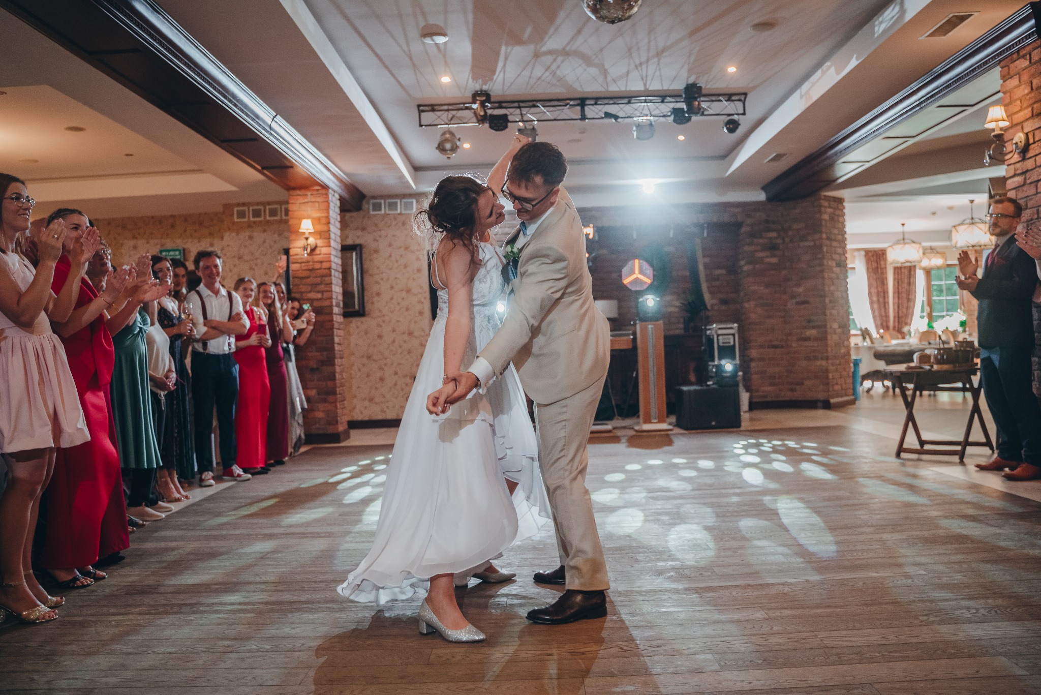 Para młoda tańczy pierwszy taniec na parkiecie, otoczona gośćmi weselnymi. W tle ceglane kolumny i dekoracje sali balowej. Oświetlenie punktowe tworzy romantyczny nastrój.