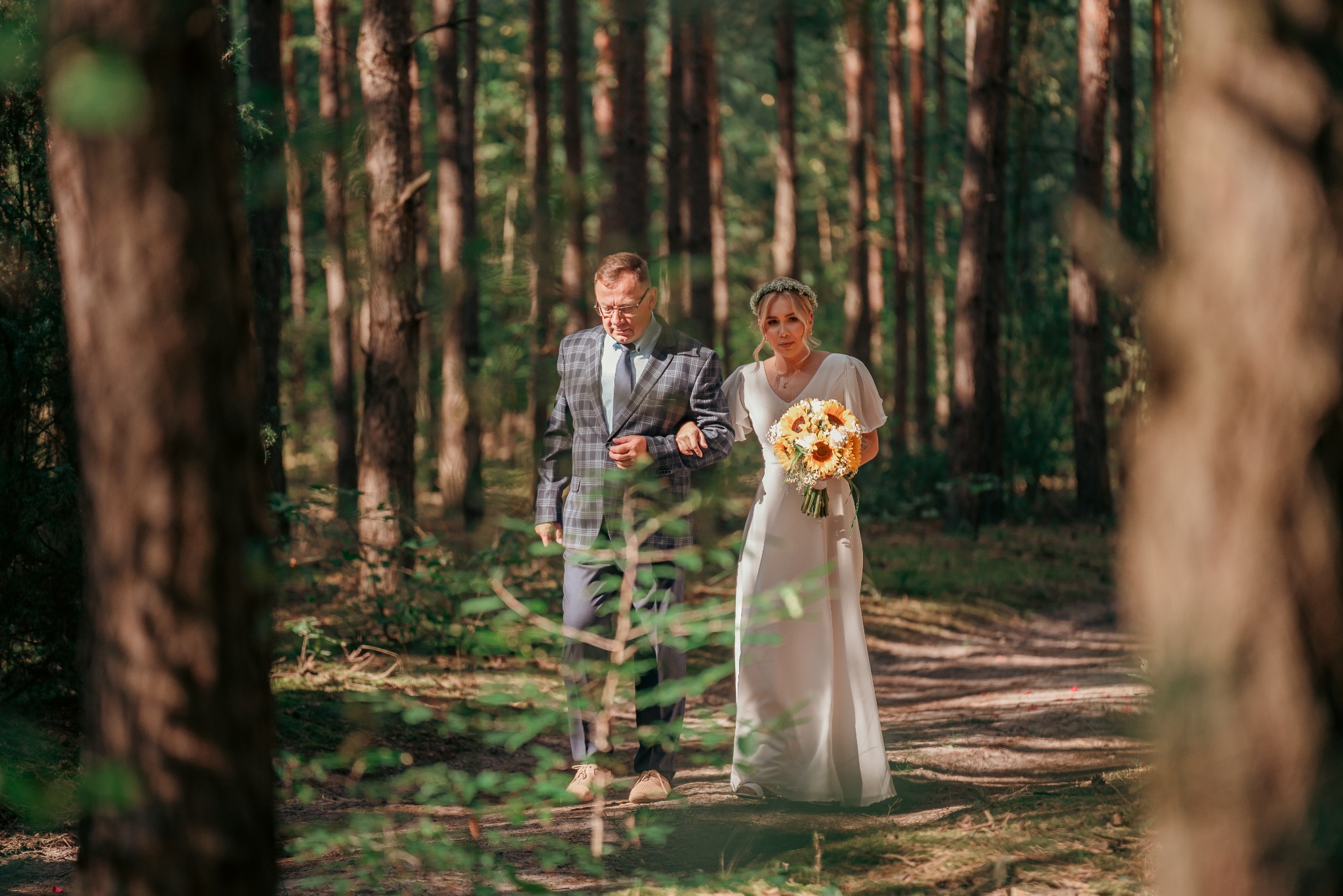 Panna młoda w zwiewnej sukni i wianku z ojcem idą leśną ścieżką, ona trzyma bukiet słoneczników. Ujęcie przez gałęzie drzew.
