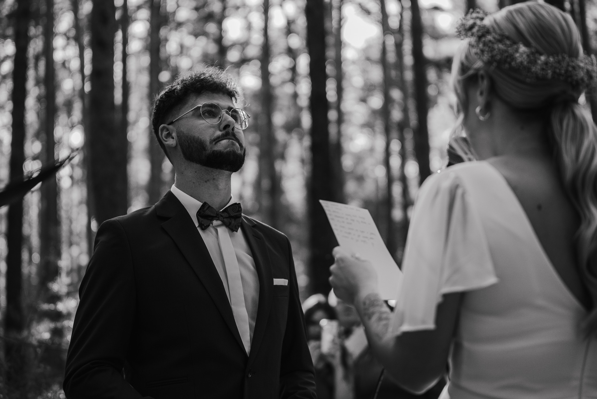 Czarno-białe zdjęcie pana młodego w garniturze i muszce, słuchającego przysięgi małżeńskiej w lesie, z widoczną panną młodą z tyłu.