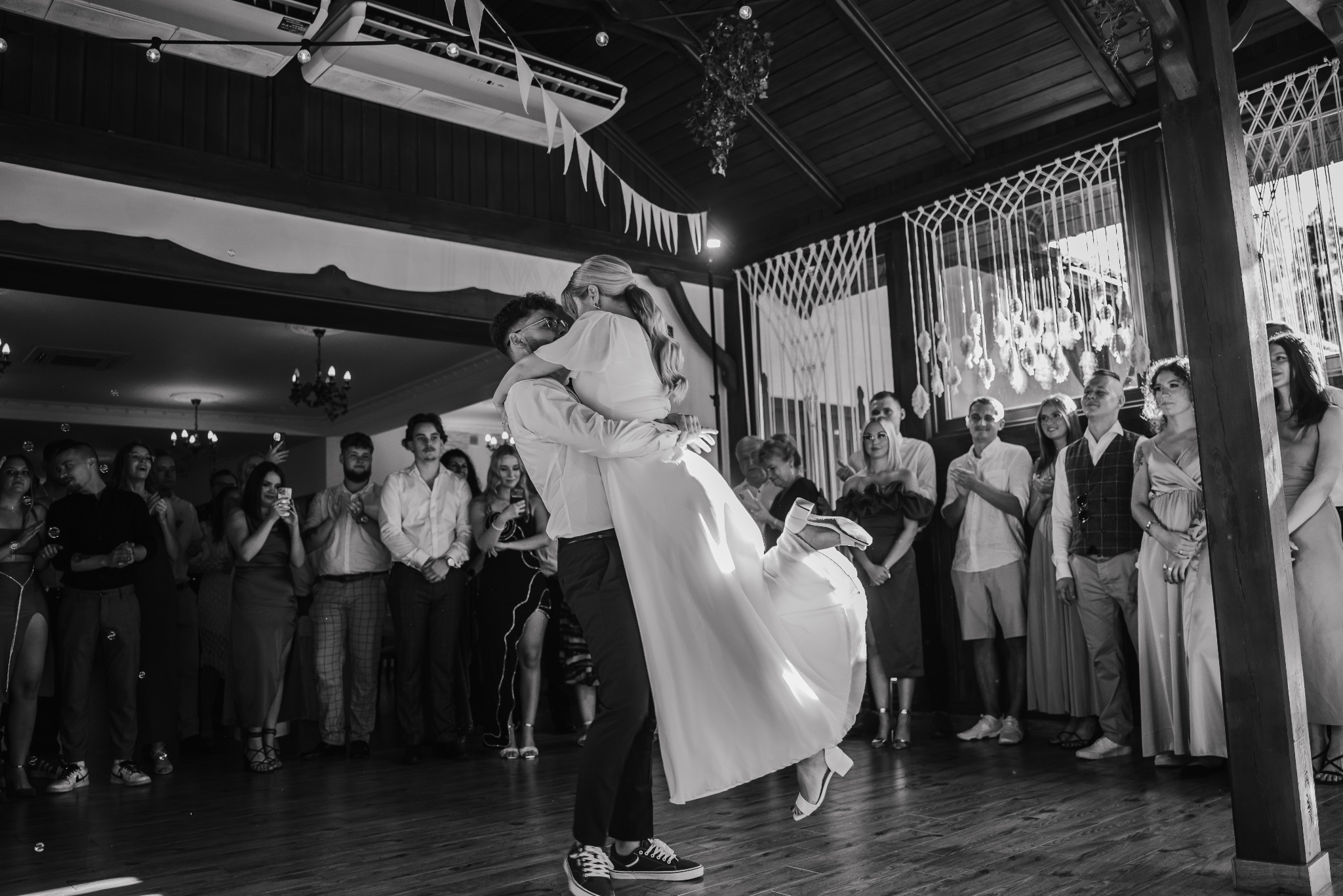 Czarno-białe zdjęcie nowożeńców podczas pierwszego tańca, unoszonych przez pana młodego na tle gości weselnych i dekoracji sali.
