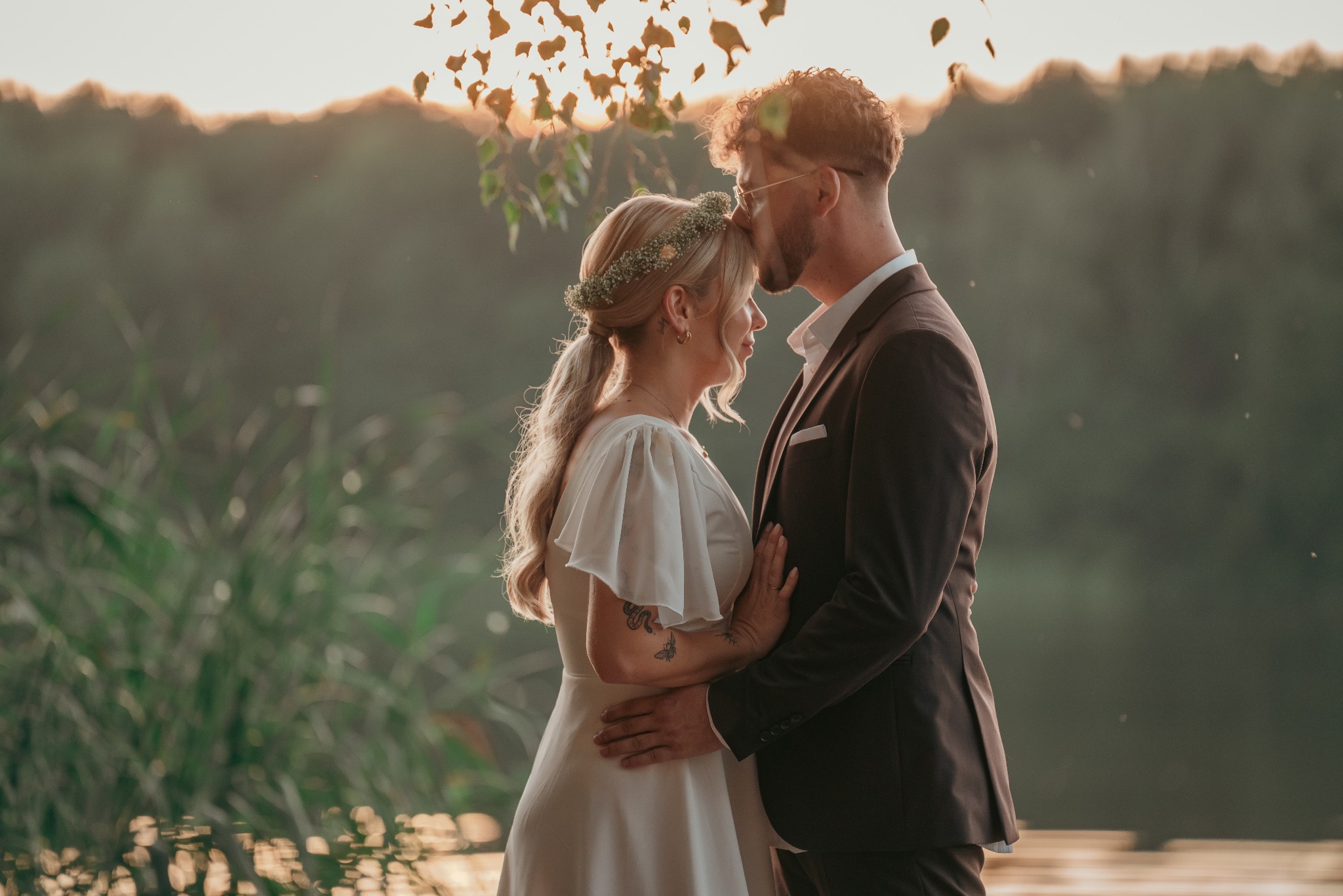 Para młoda w objęciach na tle jeziora o zachodzie słońca. Panna młoda w wianku i sukni, pan młody w garniturze. Romantyczna atmosfera, ciepłe światło.