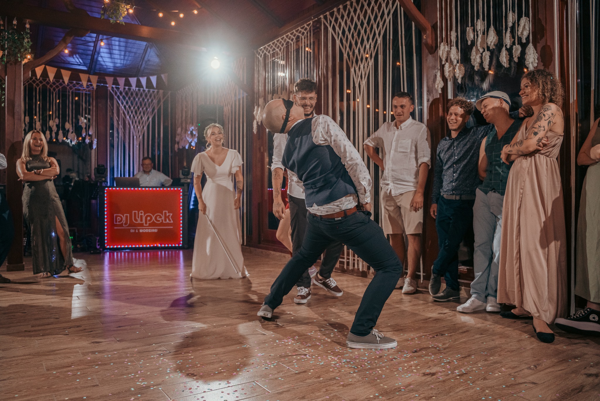 Radosna zabawa weselna w Zielonej Górze: Pan młody z zawiązanymi oczami próbuje złapać kogoś z gości na parkiecie, w tle uśmiechnięta para młoda i DJ.