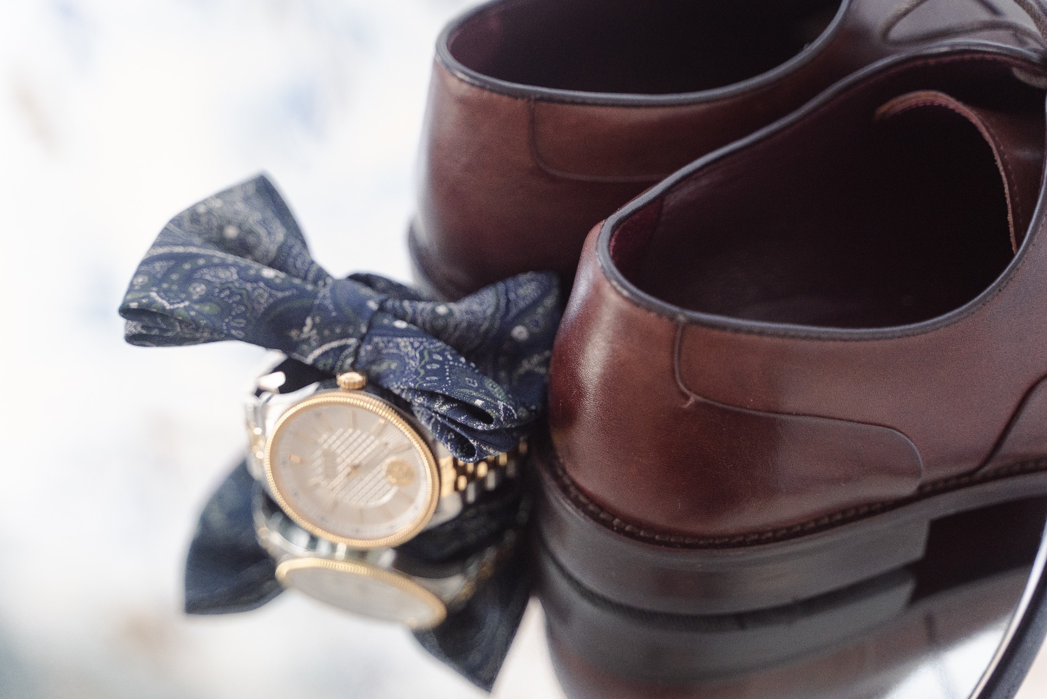 Brązowe, skórzane buty męskie, zegarek z białą tarczą i wzorzysta mucha na lustrzanej powierzchni. Przygotowania do ślubu, detale.