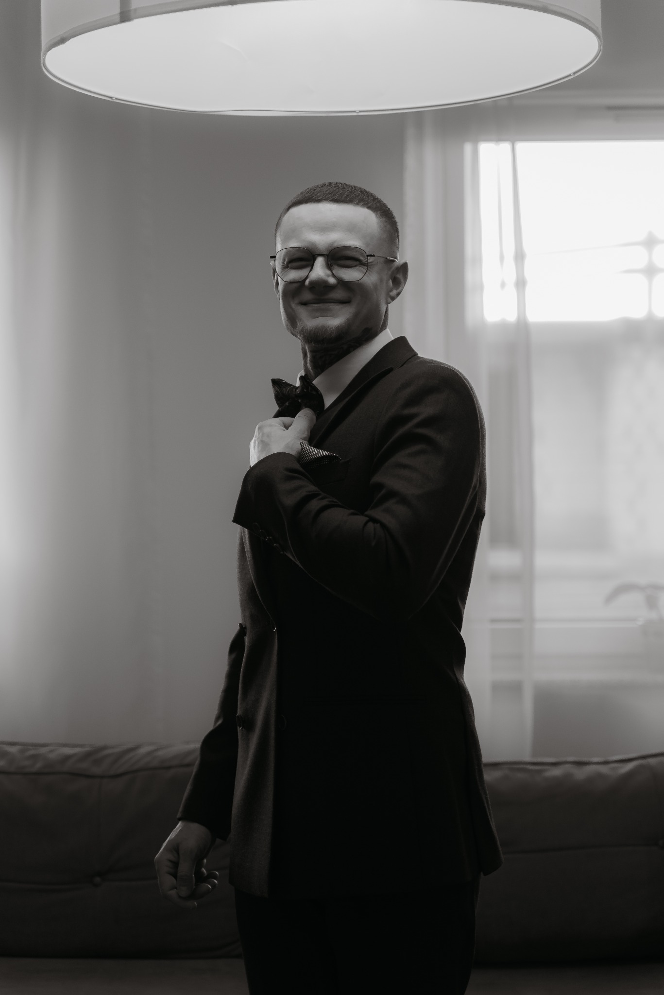 Czarno-białe zdjęcie uśmiechniętego mężczyzny w garniturze i okularach, poprawiającego muchę. W tle okno i lampa, tworzące subtelne oświetlenie.