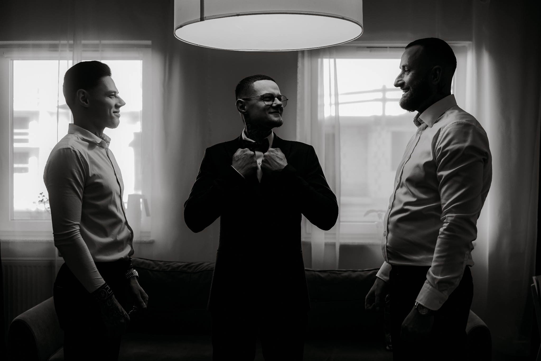 Czarno-białe zdjęcie trzech mężczyzn w koszulach, jeden w garniturze, poprawiający muchę, w jasnym wnętrzu z oknami, uśmiechnięci, przygotowania do uroczystości.