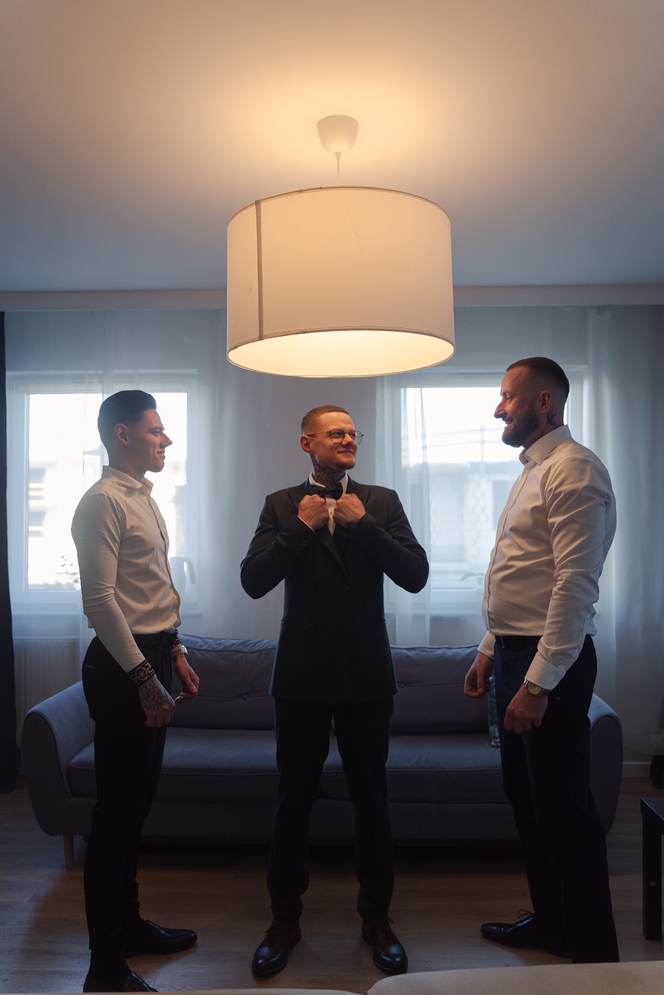 Trzech mężczyzn w eleganckich strojach, jeden poprawia muchę, w jasnym wnętrzu z szarą sofą i lampą. Ujęcie z dołu podkreśla wysokość pomieszczenia.
