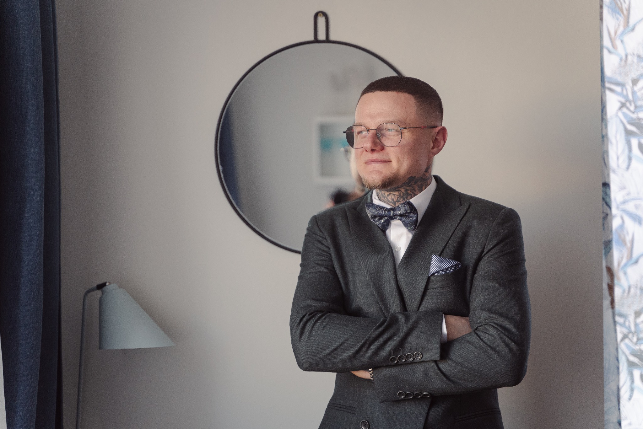 Elegancki mężczyzna w garniturze i muszce, z tatuażami na szyi, stoi na tle lustra i lampy, spoglądając w bok. Ujęcie portretowe, stonowana kolorystyka.