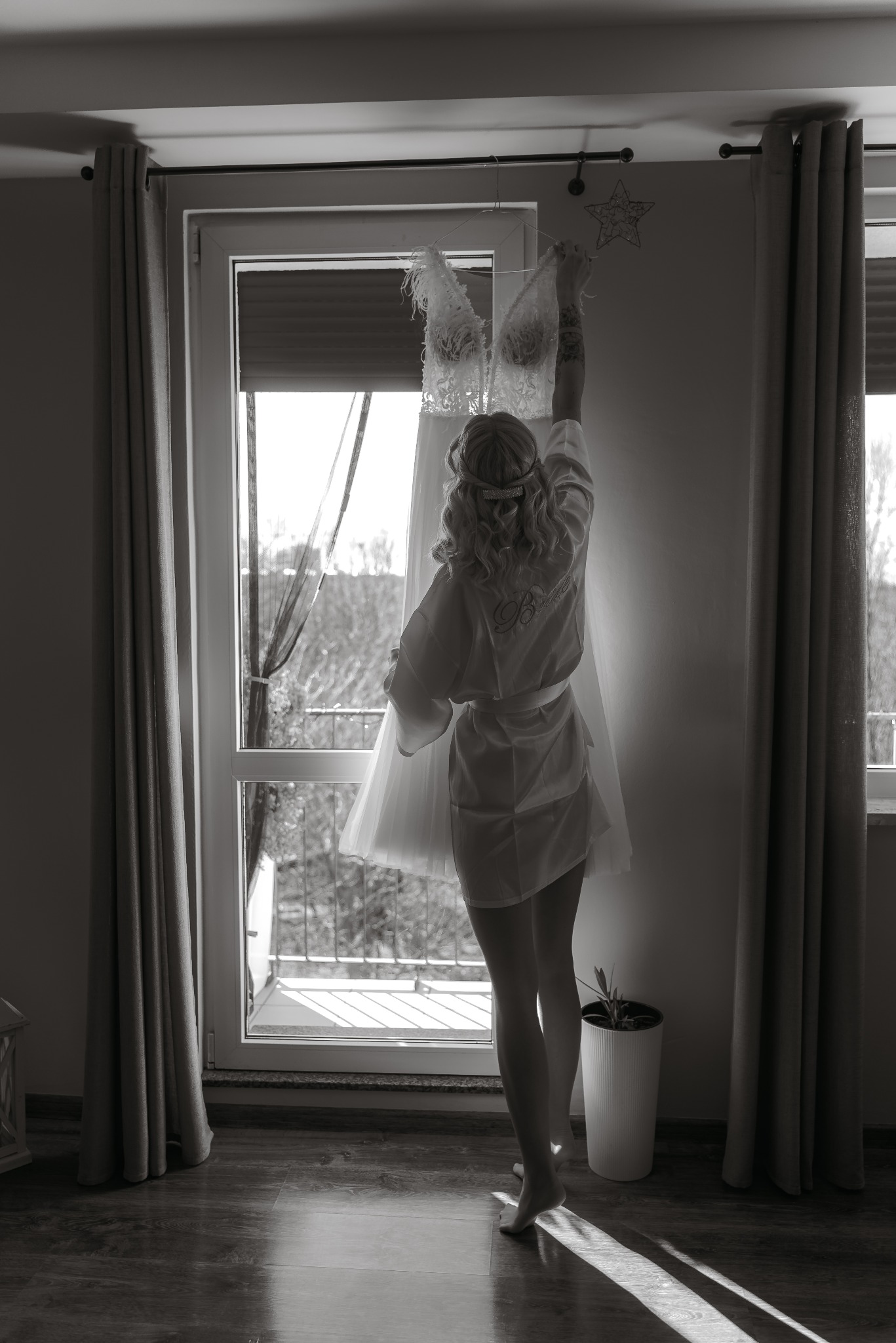 Panna młoda w szlafroku wiesza suknię ślubną na oknie. Ujęcie zza pleców, czarno-białe, z widocznym tatuażem na ramieniu i dekoracją w kształcie gwiazdy na ścianie.