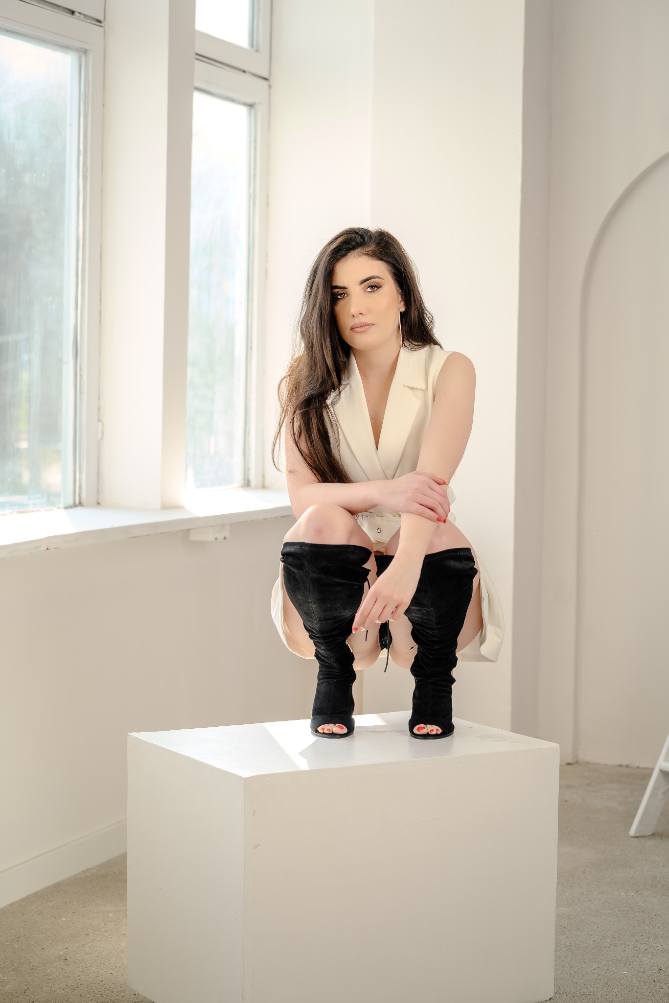 Kobieta w beżowej sukience i czarnych kozakach pozuje, siedząc na białym postumencie na tle jasnego okna. Minimalistyczna sceneria, naturalne światło, subtelny makijaż.
