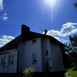 K&M Elewacje - Pracownik na dachu domu podczas mycia elewacji pod silnym słońcem. Widoczne rusztowanie, ogród i fragment sąsiedniego budynku. Ujęcie z dołu.