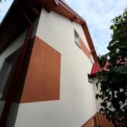 Dwukolorowa elewacja domu z kwadratowym akcentem w odcieniach brązu, widoczna z dołu, z fragmentem czerwonego dachu i rynny. Nowoczesny design.