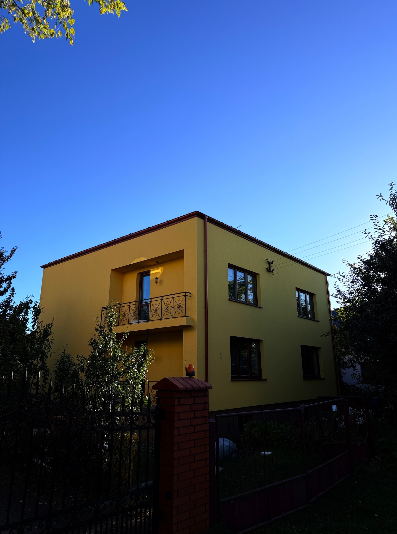 Żółty dom z balkonem i ciemną stolarką okienną, widziany z dołu pod błękitnym niebem. Ciemne ogrodzenie i zieleń drzew w dolnej części kadru.