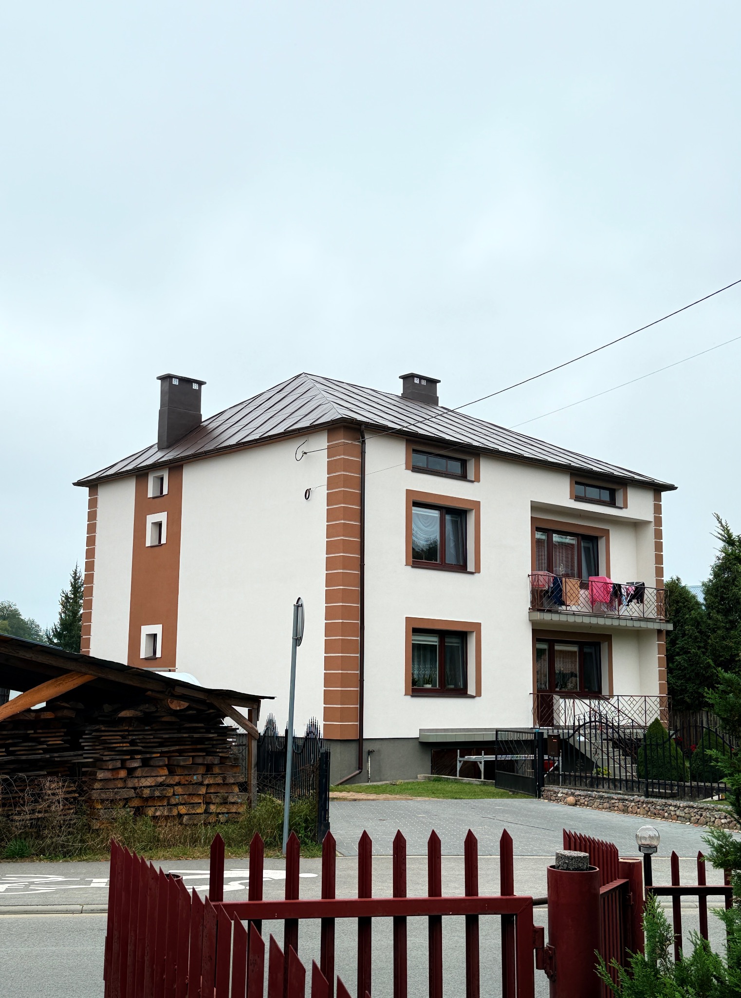 Dwupiętrowy dom z balkonem i brązowymi akcentami elewacji, widoczny zza czerwonego płotu. Na balkonie suszą się ubrania. Obok domu skład drewna pod zadaszeniem.