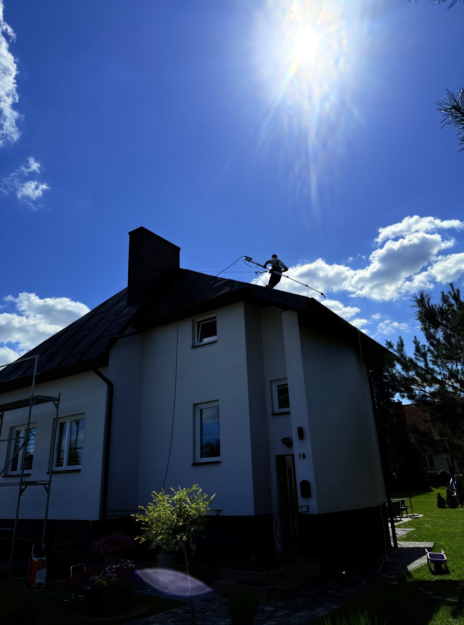 Pracownik na dachu domu podczas mycia elewacji pod silnym słońcem. Widoczne rusztowanie, ogród i fragment sąsiedniego budynku. Ujęcie z dołu.