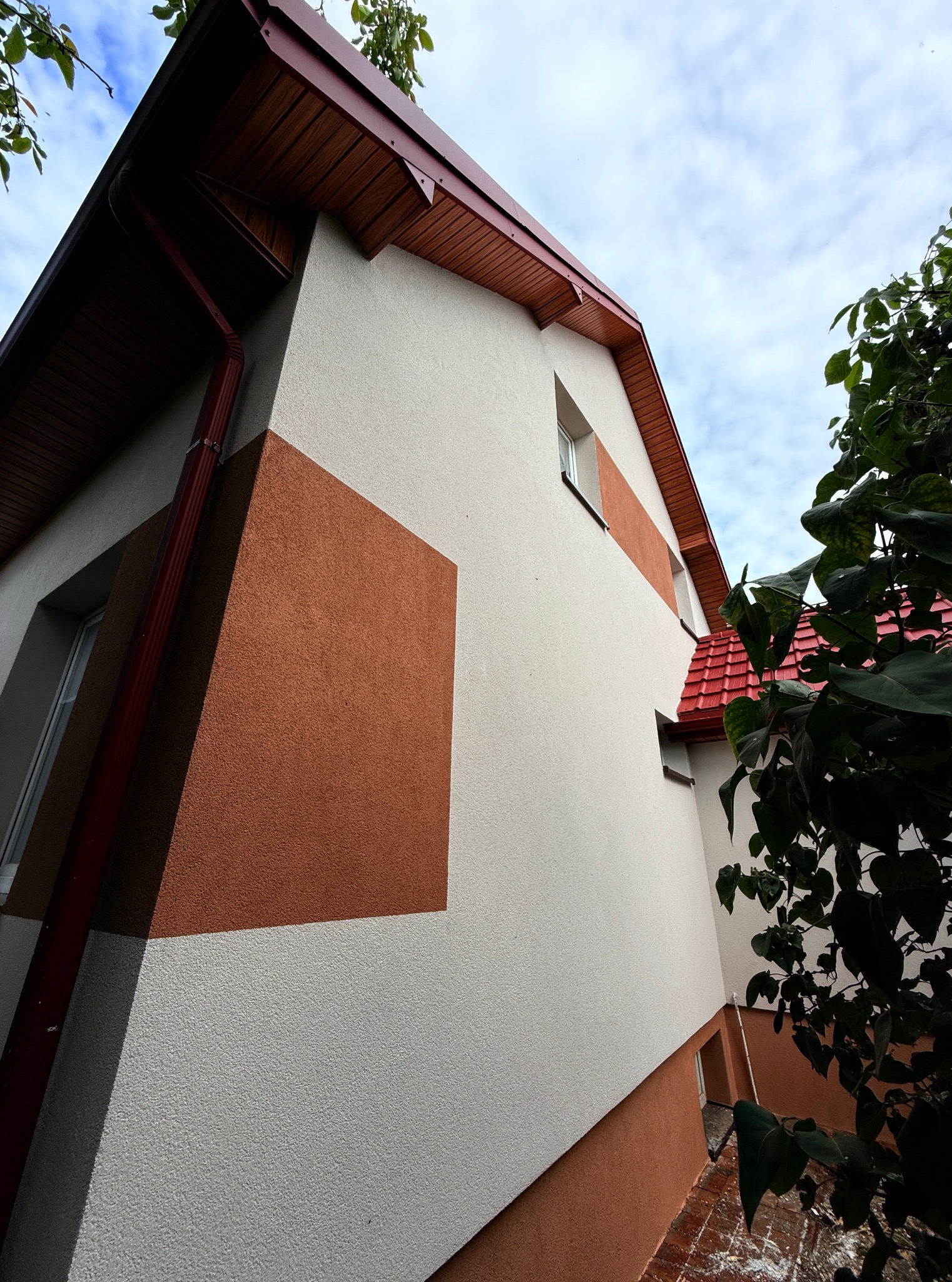 Dwukolorowa elewacja domu z kwadratowym akcentem w odcieniach brązu, widoczna z dołu, z fragmentem czerwonego dachu i rynny. Nowoczesny design.