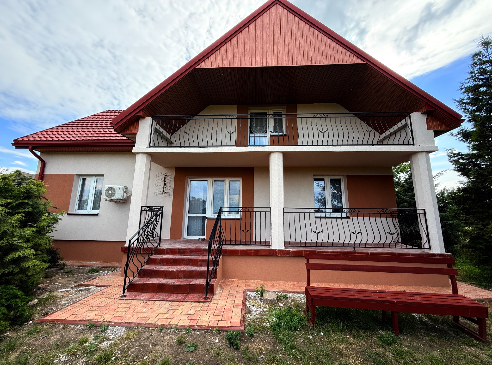 Dwukondygnacyjny dom z balkonem i czerwonym dachem, widoczny z poziomu gruntu. Schody wejściowe z czerwoną kostką brukową i metalową balustradą. Na ścianie klimatyzator.