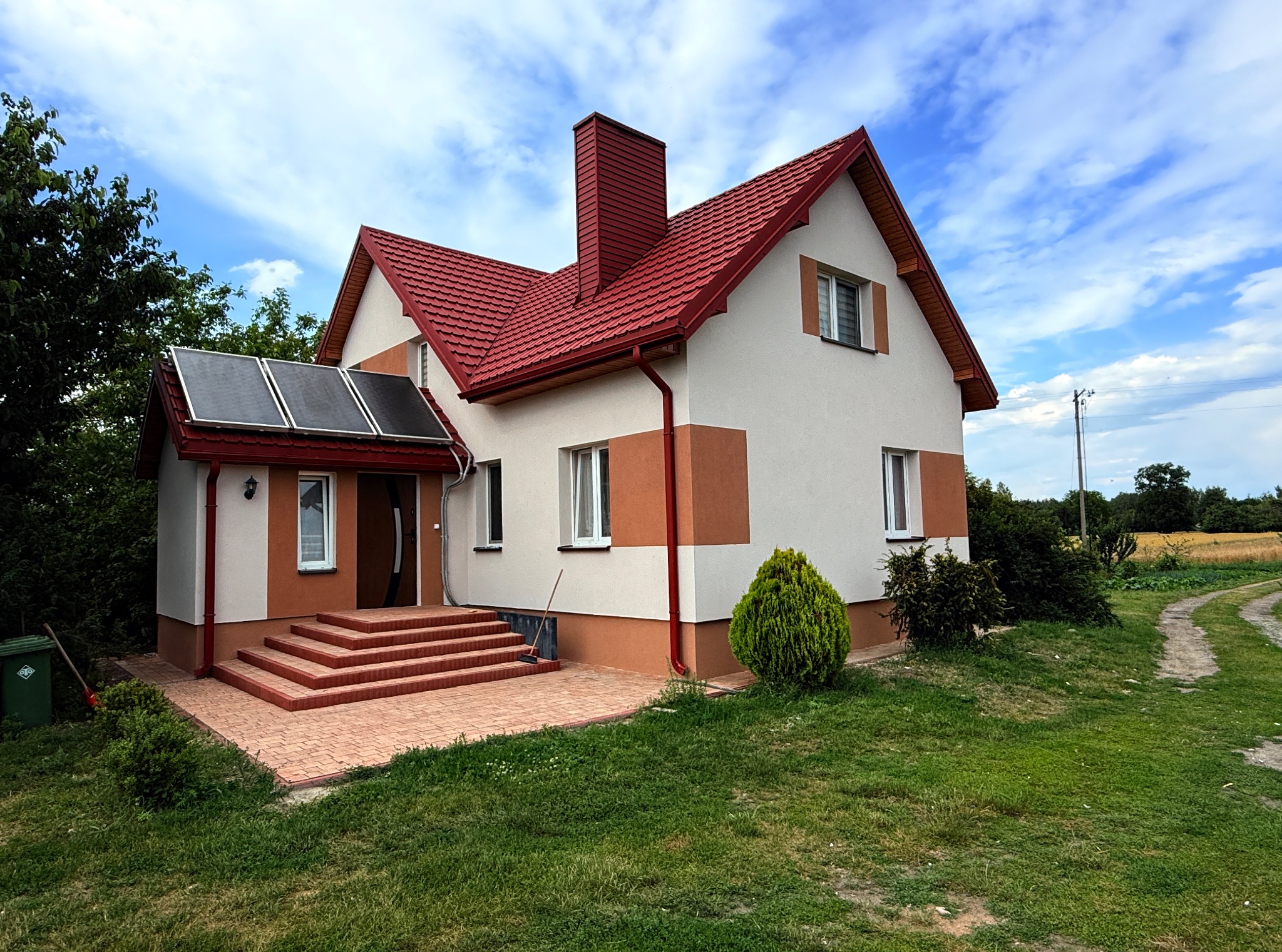Dwukondygnacyjny dom z czerwonym dachem i panelami słonecznymi nad wejściem, otoczony zielonym trawnikiem i ścieżką. Widoczne elementy elewacji w dwóch odcieniach.