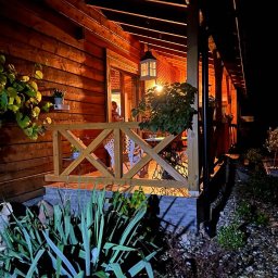 Arkadiusz Wojciech - Oświetlony, drewniany taras z balustradą i roślinami w nocy. Lampa wisząca nad stołem i krzesłami. Widok na wnętrze domu przez otwarte drzwi.