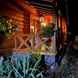 Oświetlony, drewniany taras z balustradą i roślinami w nocy. Lampa wisząca nad stołem i krzesłami. Widok na wnętrze domu przez otwarte drzwi.