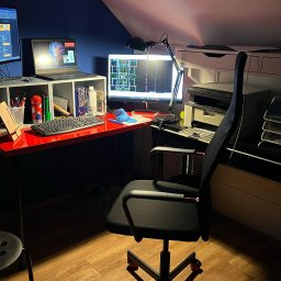 Arkadiusz Wojciech - Ergonomiczne biuro w pokoju na poddaszu: czerwone biurko, monitory z planami, laptop, lampka, krzesło biurowe. Projektowanie wnętrz biura domowego.