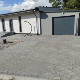 Szara kostka brukowa ułożona na podjeździe przed nowoczesnym domem z garażem. Widoczne elementy architektury i fragment ogrodu.