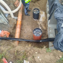 Rob-Bruk usługi brukarskie i budowlane - Wykop z ułożonymi rurami drenarskimi, kablami i studzienkami. Widoczny pracownik w kamizelce odblaskowej. Prace ziemne w trakcie realizacji.