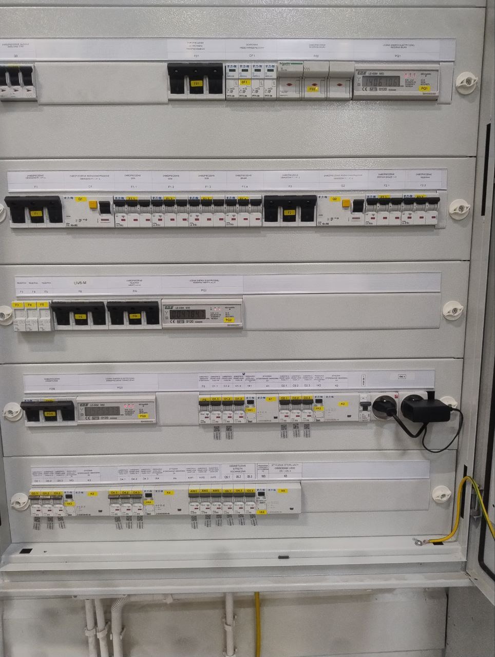 Szara skrzynka elektryczna z bezpiecznikami, opisana etykietami i oznaczeniami, widoczne przewody. Instalacja elektryczna wewnątrz budynku.