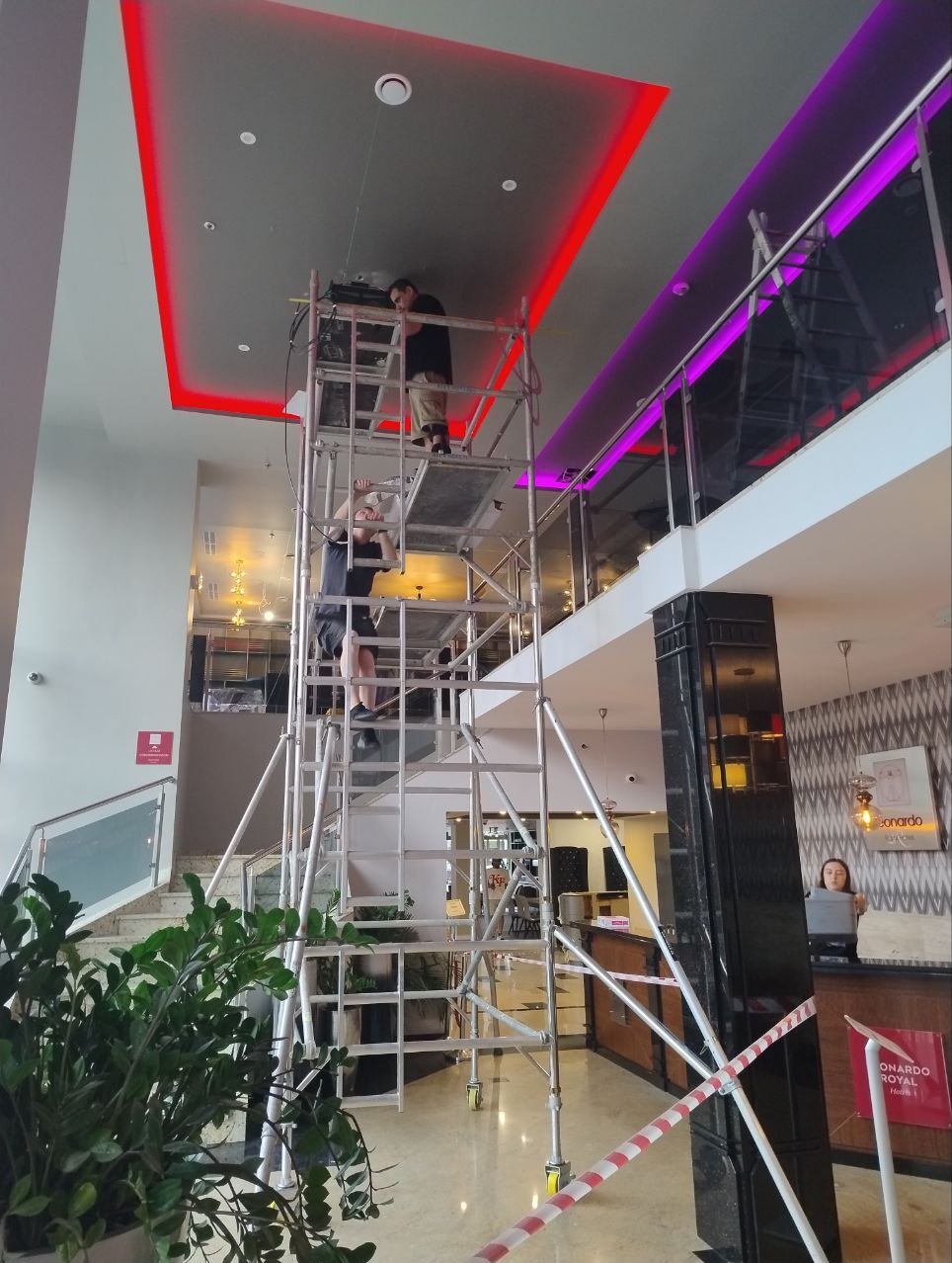 Dwóch monterów na rusztowaniu instaluje oświetlenie LED w suficie holu hotelowego. Widoczne czerwone i fioletowe pasy świetlne, recepcja w tle.