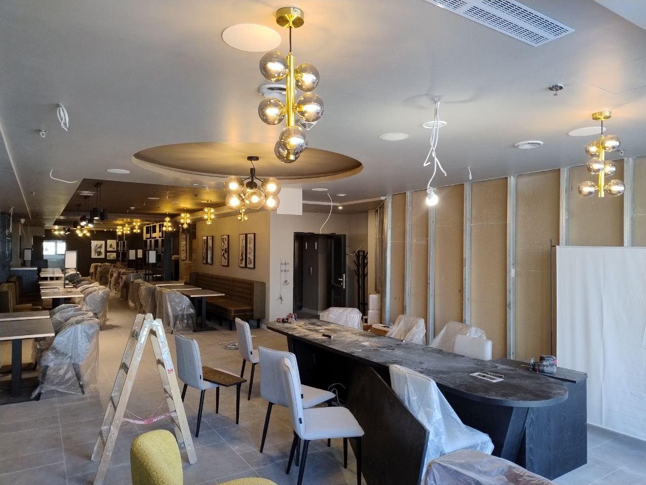 Wnętrze lokalu w trakcie wykańczania z zamontowanym oświetleniem: wiszące lampy z kulistymi kloszami. Widoczne rusztowanie i zabezpieczone meble.