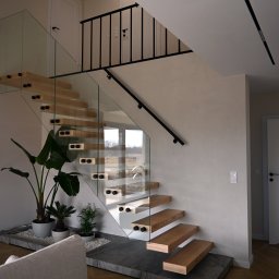 STOLARNIA MODŁA SPÓŁKA Z OGRANICZONĄ ODPOWIEDZIALNOŚCIĄ - Nowoczesne schody z drewnianymi stopniami i szklaną balustradą, wspornikowe, z czarnymi elementami mocującymi, u podstawy rośliny doniczkowe na kamiennym podłożu.