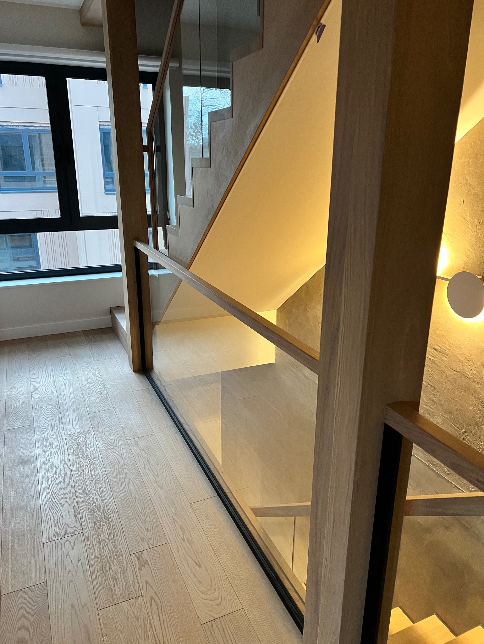 Nowoczesne schody drewniane z przeszkloną balustradą, oświetlone ciepłym światłem, widoczne z perspektywy podłogi z jasnego drewna. Minimalistyczny design.