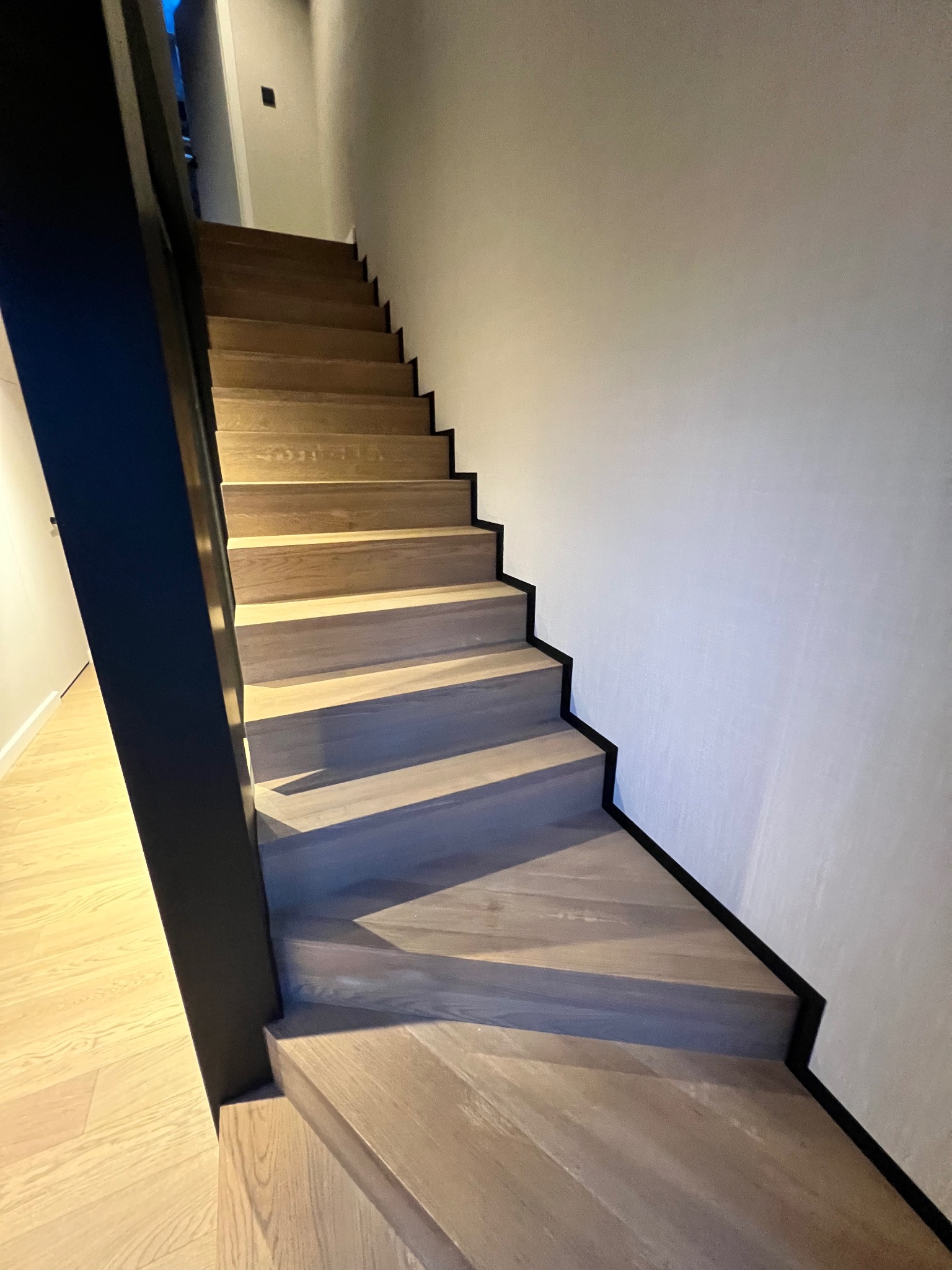 Nowoczesne schody drewniane z czarną balustradą i listwami przypodłogowymi, widok z dołu, jasne drewno, minimalistyczny design, oświetlenie górne.