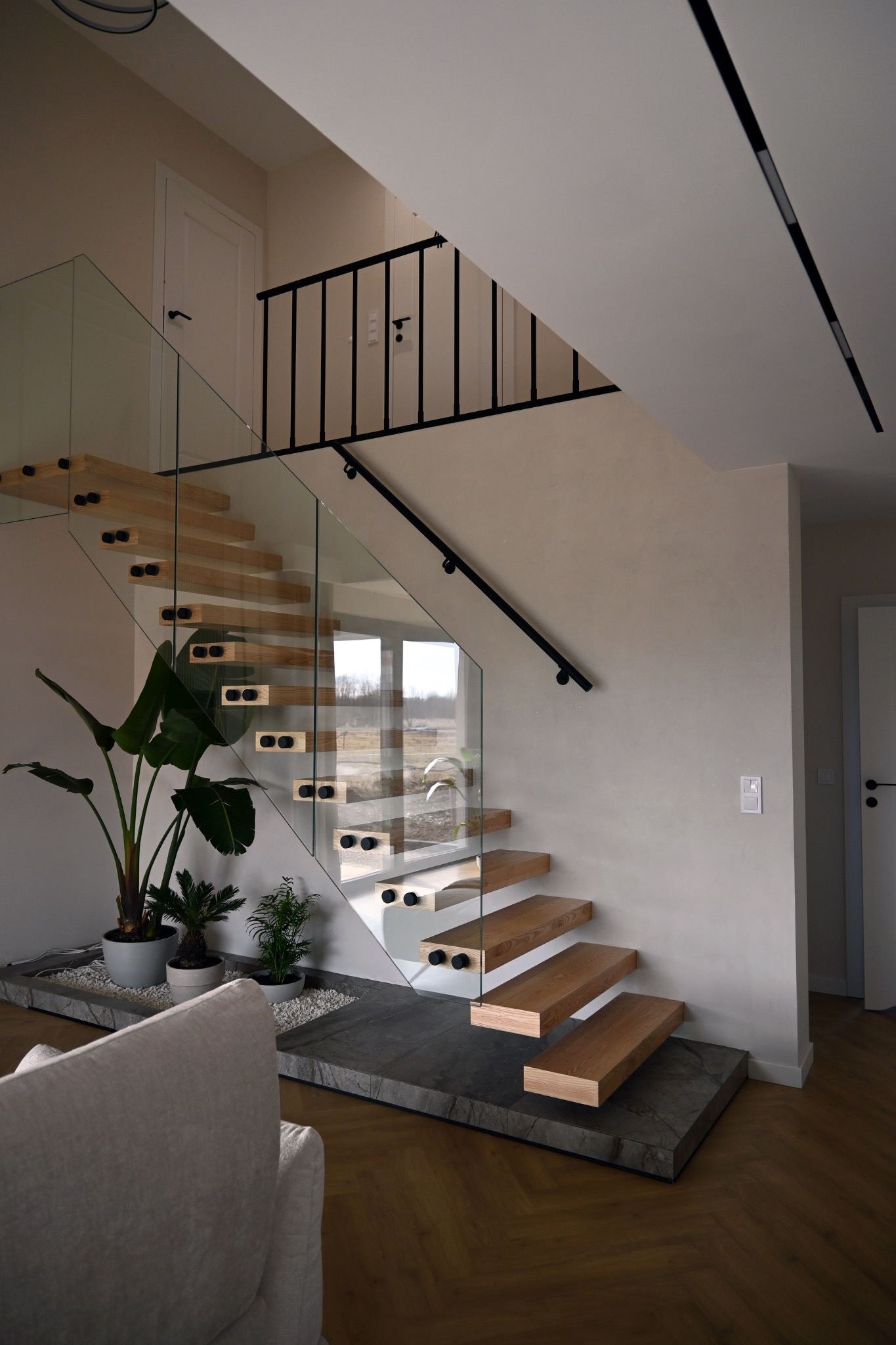 Nowoczesne schody z drewnianymi stopniami i szklaną balustradą, wspornikowe, z czarnymi elementami mocującymi, u podstawy rośliny doniczkowe na kamiennym podłożu.