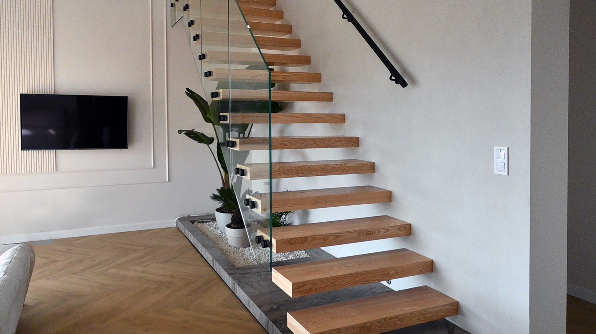 Nowoczesne schody drewniane z balustradą szklaną i czarną poręczą, stopnie wspornikowe, minimalistyczny design, jasne wnętrze z parkietem w jodełkę.