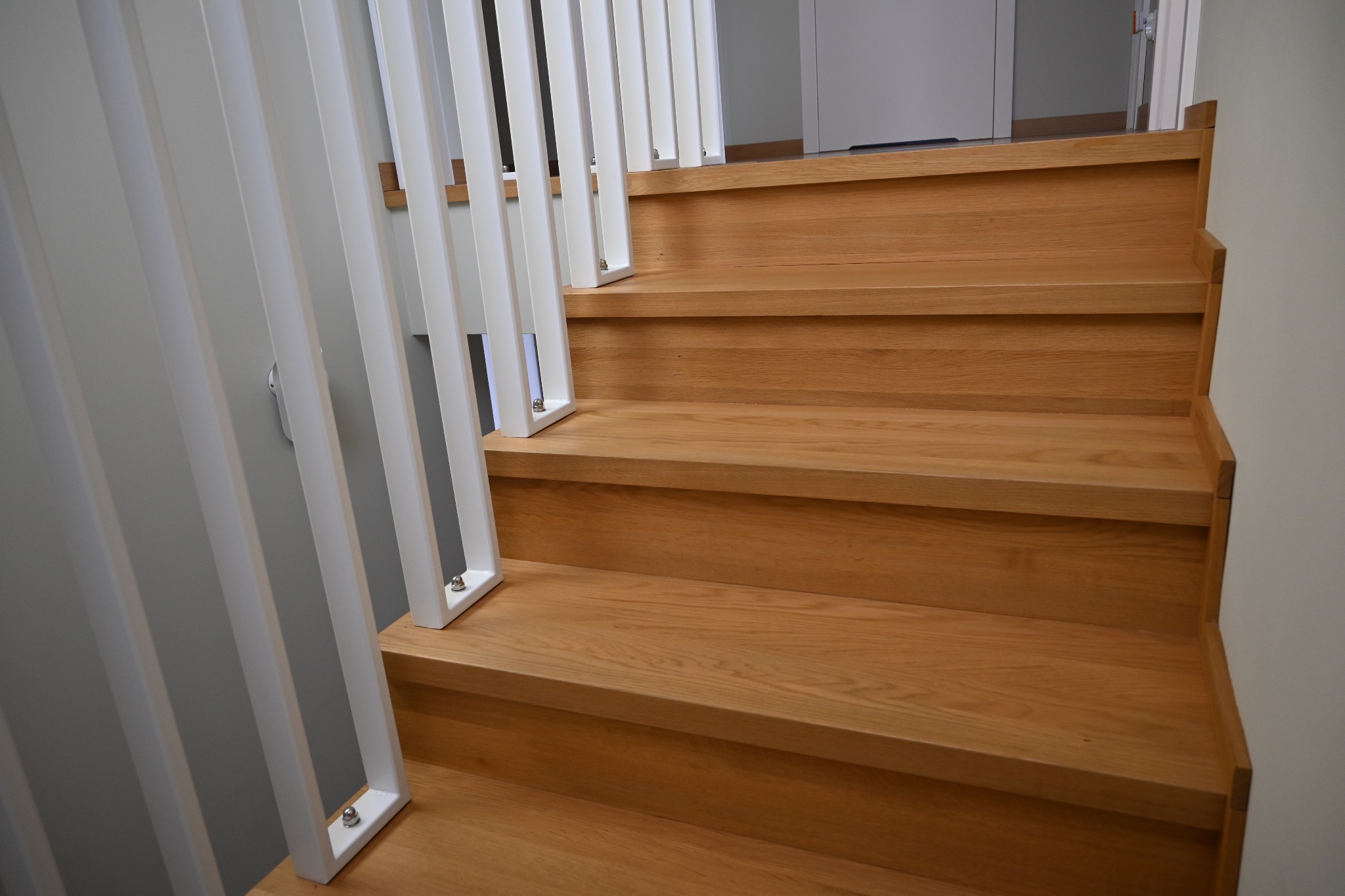 Jasne, dębowe schody z białą, nowoczesną balustradą. Minimalistyczny design, widoczne detale łączeń. Naturalne światło podkreśla strukturę drewna.