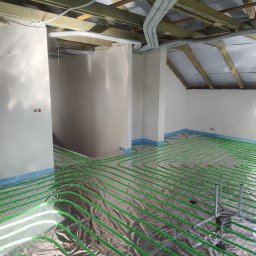 Gaga - Instalacja ogrzewania podłogowego z zielonych rur na folii odblaskowej w surowym wnętrzu poddasza. Widoczne rury wentylacyjne pod drewnianą konstrukcją dachu.