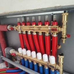 Gaga - Rozdzielacz ogrzewania podłogowego z czerwonymi i niebieskimi rurami, zamontowany w szarej szafce. Widoczne napisy 'SAU VC', 'SIŁOWNIA' i 'OUT'.