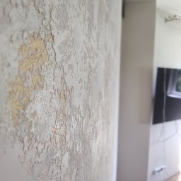 Gaga - Dekoracyjna ściana z efektem strukturalnym w odcieniach bieli i złota, widoczna z bliska, z fragmentem wnętrza w tle. Unikalna faktura i oświetlenie podkreślają detale.