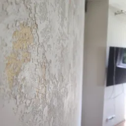 Dekoracyjna ściana z efektem strukturalnym w odcieniach bieli i złota, widoczna z bliska, z fragmentem wnętrza w tle. Unikalna faktura i oświetlenie podkreślają detale.