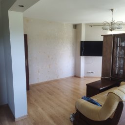 Gaga - Jasne wnętrze salonu po malowaniu ścian z dekoracyjną fakturą. Widoczny fragment sofy, drewniana podłoga i wiszący telewizor. Oświetlenie sufitowe.