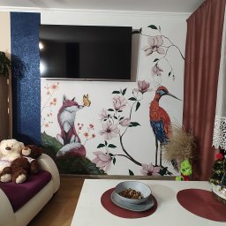 Gaga - Pokój z dekoracyjną ścianą: malunek lisa z motylem i żurawia wśród magnolii. Telewizor na ścianie, sofa z pluszakami i stół z dekoracjami.