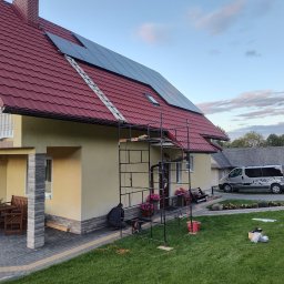 Gaga - Montaż paneli fotowoltaicznych na dachu domu z czerwoną dachówką. Widoczne rusztowanie, drabina i częściowo sylwetka instalatora. Na trawniku narzędzia i akcesoria.