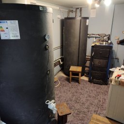 Gaga - Dwa czarne zbiorniki akumulacyjne ciepła w kotłowni, widoczne rury i armatura, mały drewniany stołek i piec węglowy w tle, oświetlenie sufitowe.