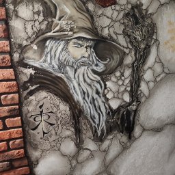Gaga - Artystyczne malowidło na ścianie z motywem czarodzieja w kapeluszu, z brodą i laską, obok fragmentu ściany z cegły. U góry oko Saurona. Styl fantasy, unikalna faktura.